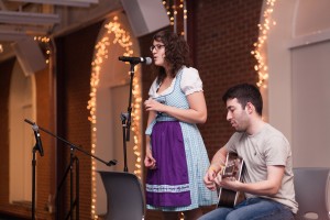 A person sining into a microphone while another is sitting and playing the guitar.